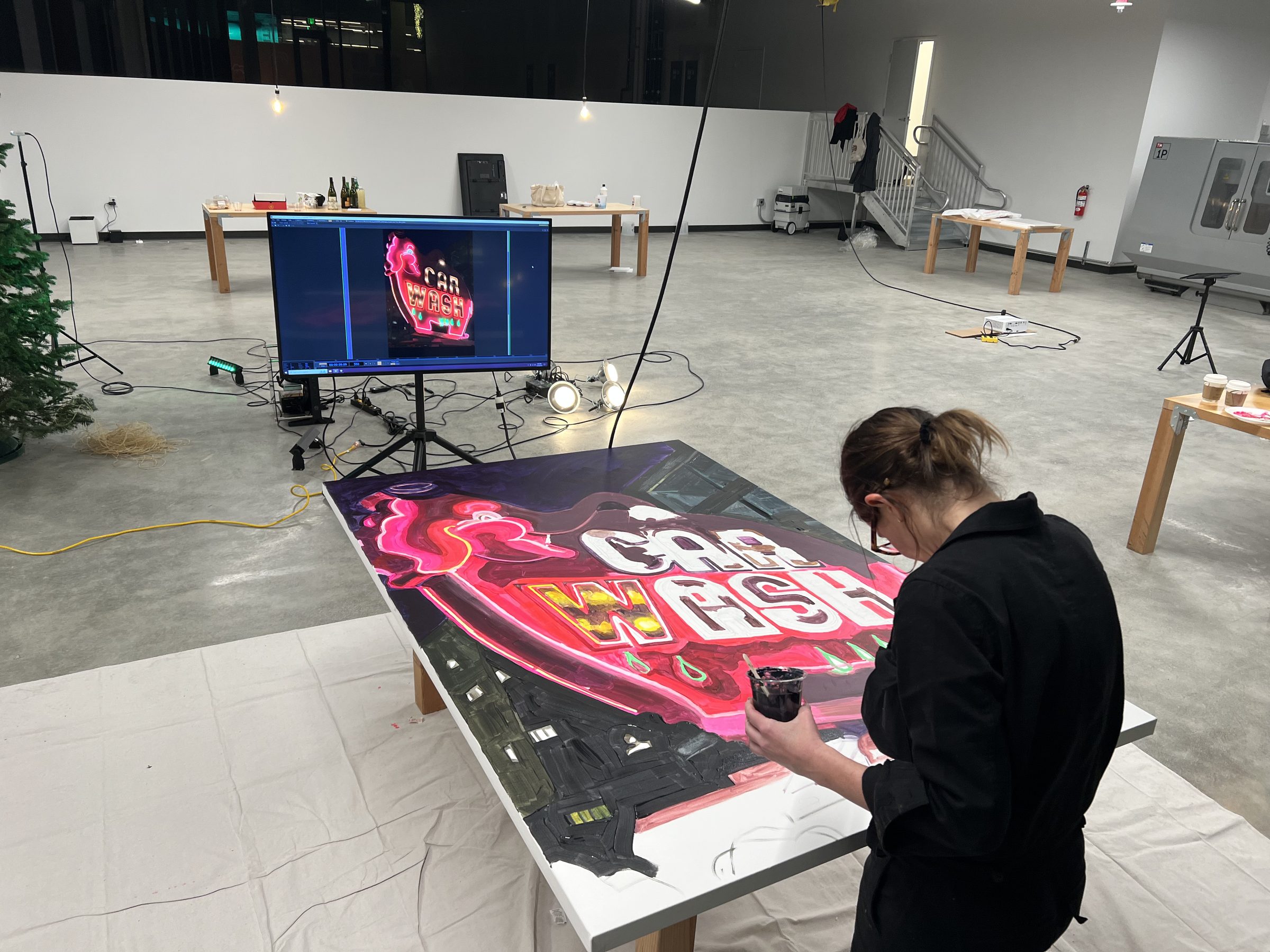 Jacqueline Goldberg at the studio table painting the Pink Elephant panel, with the original neon sign visible on the reference monitor at left.