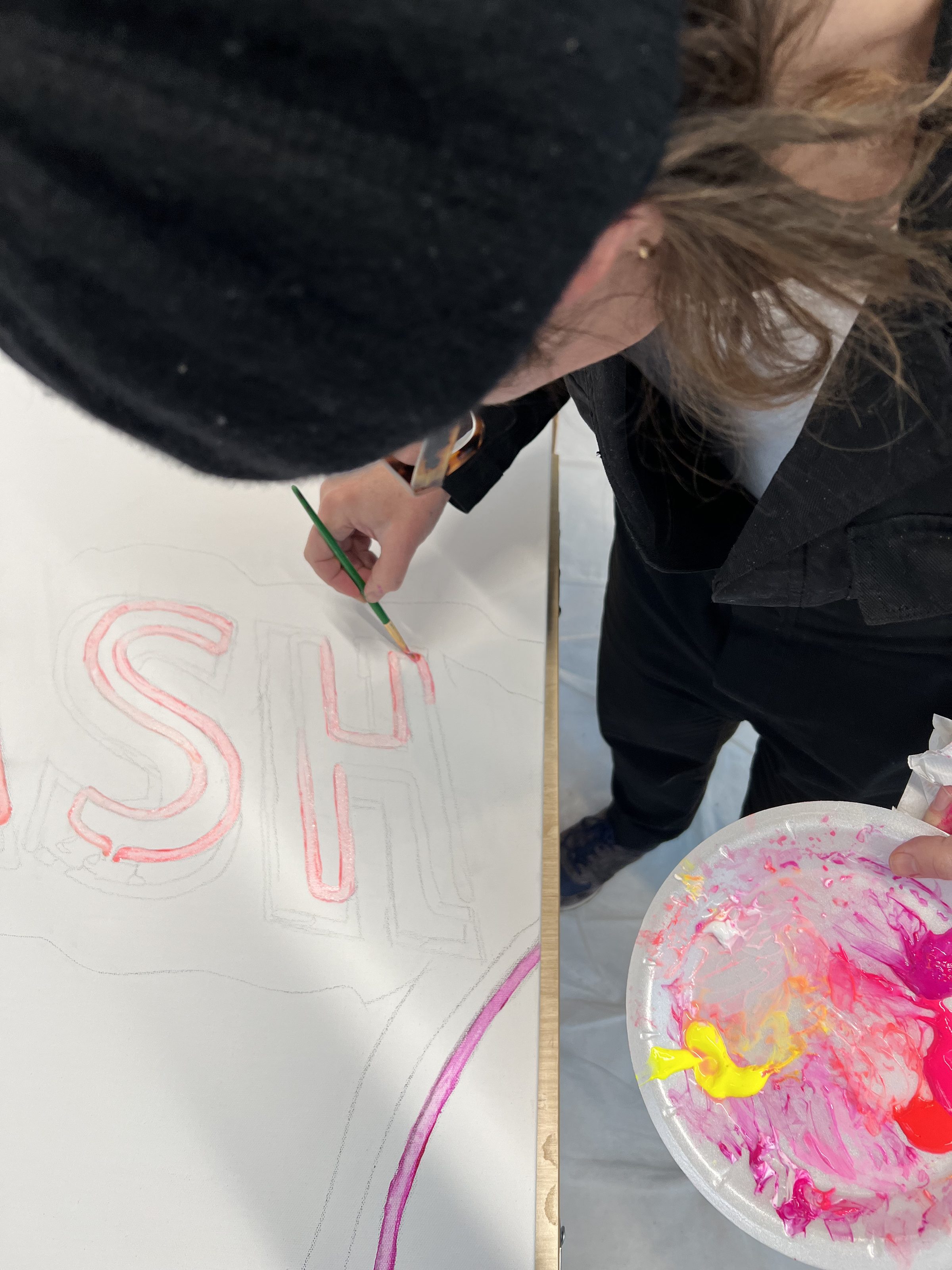 Close overhead view of hands at the canvas, palette of pinks and yellow, brush poised over the chalked lettering.