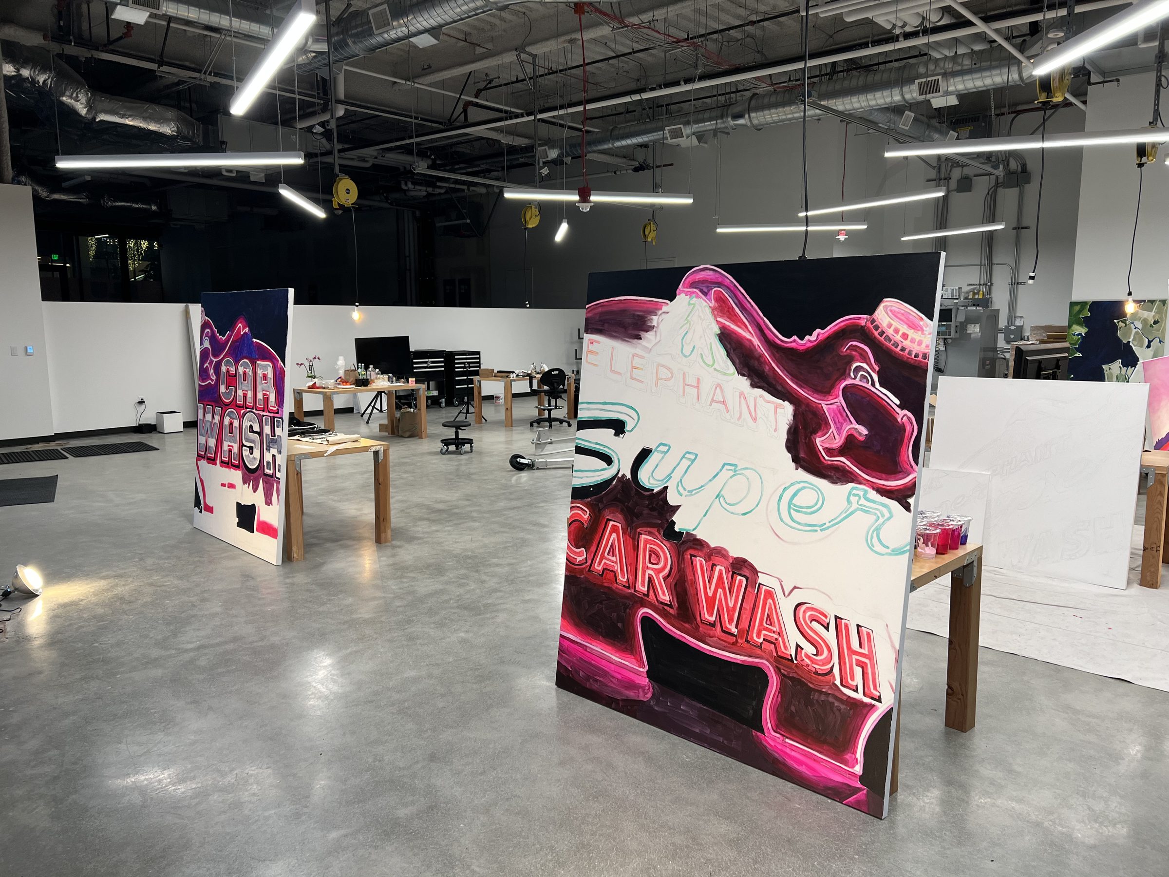 Two paintings standing in the still-empty Ground Floor Studio — CAR WASH at left, the Pink Elephant Super Car Wash at centre — overhead lights raking across.